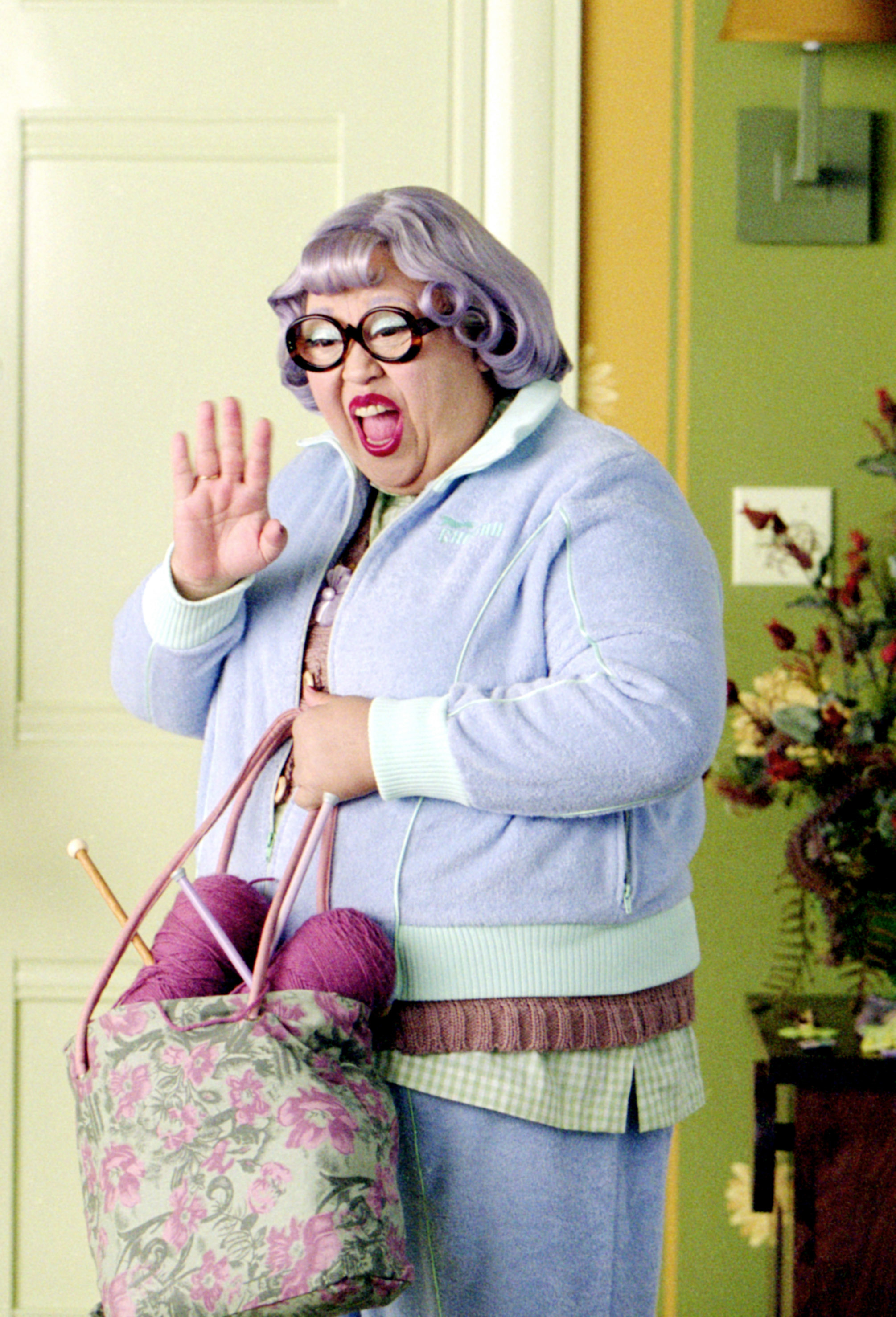 in a scene, Amy in a sweater and stylized wig and waving with a bag and yarn