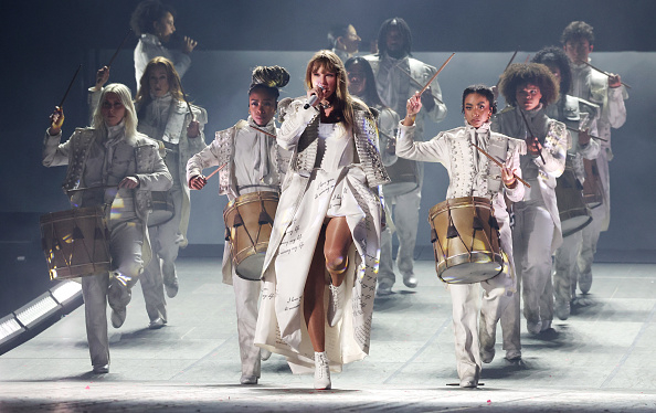 Performer in an elaborate coat with backup dancers in uniform, on stage with drums