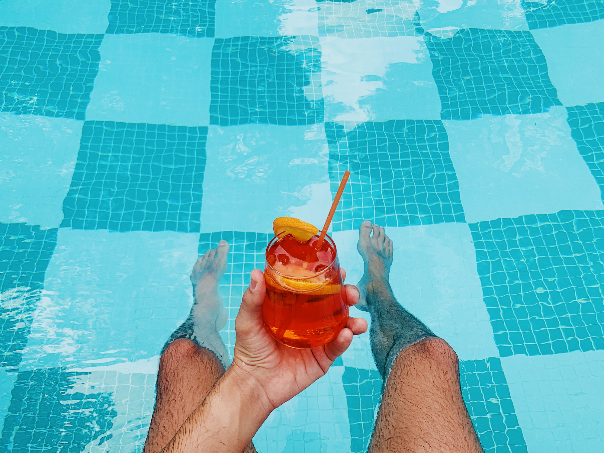 Person holding a drink by a pool, legs submerged in water, showcasing leisure and relaxation