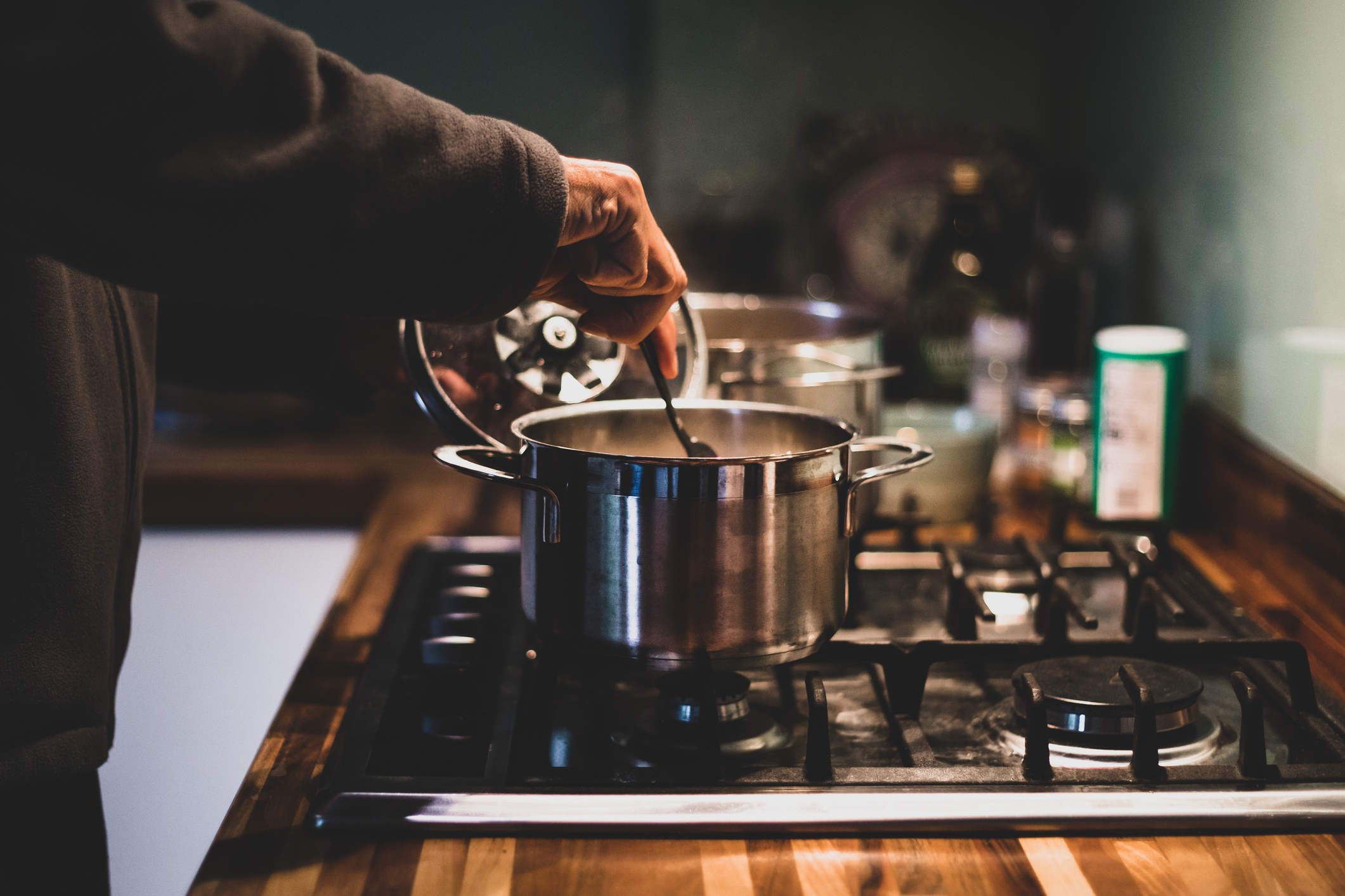 Person cooking, lifting lid off pot on stove. Kitchen items in background