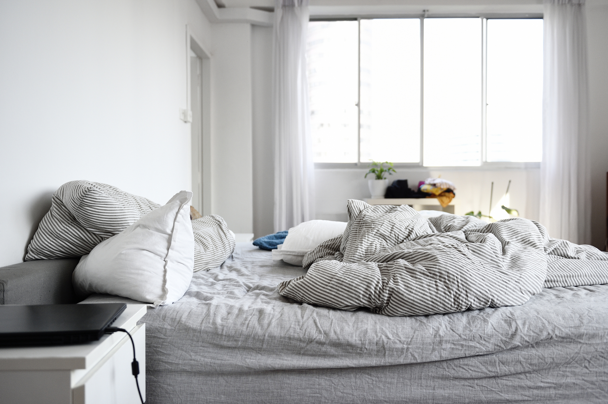 Unmade bed with striped bedding and multiple pillows in a bright room, small table and plant near window