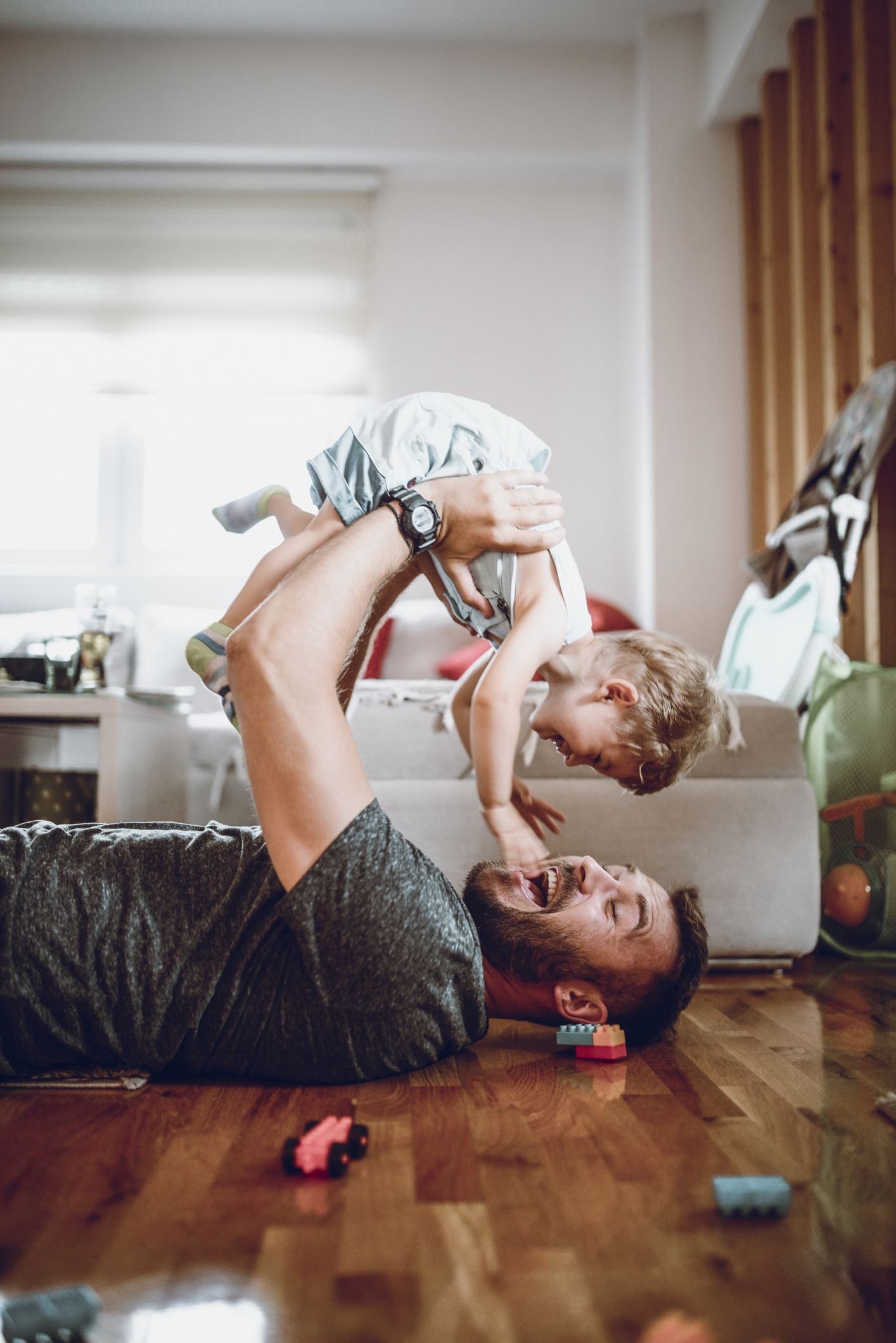 Adult playfully lifts a child while lying on the floor indoors, both laughing