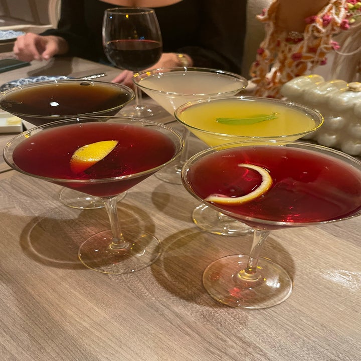 Four variously garnished cocktails on a table, with people in the background