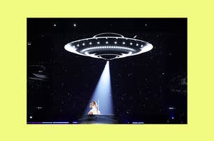 Performer on stage under a large illuminated UFO-like structure with a spotlight and audience in the background