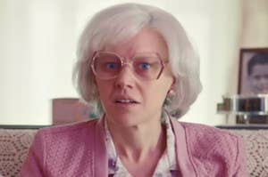 Woman in character with wig and glasses, looking surprised, in a room with a photo in the background