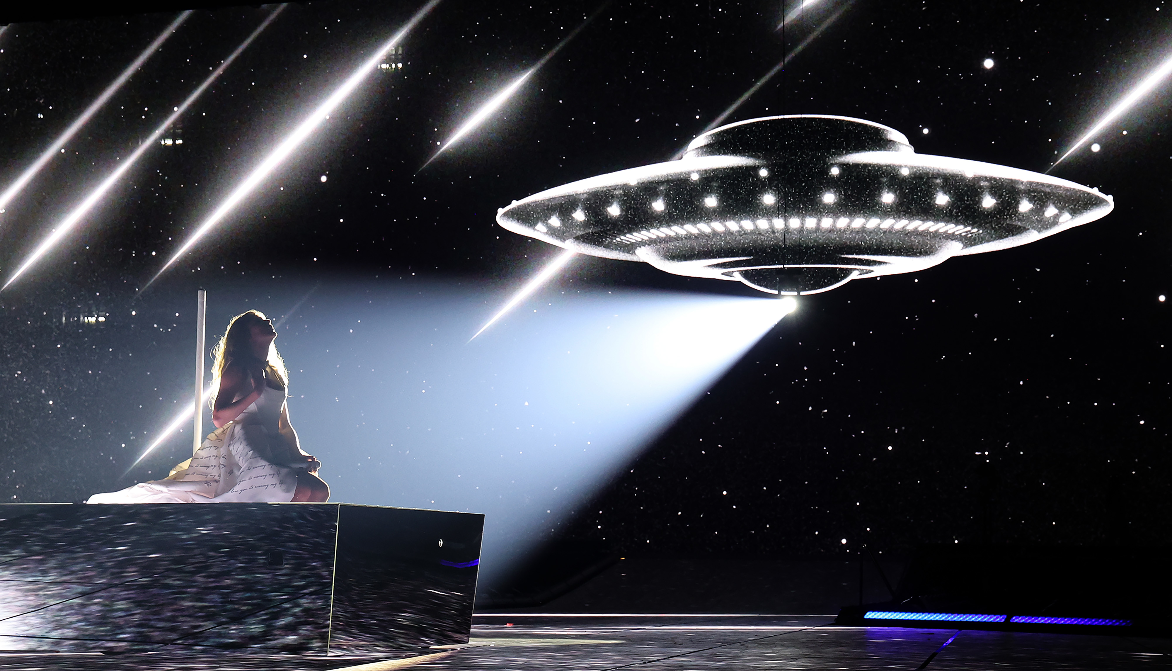Taylor Swift sits on stage under a spotlight with a UFO prop hovering above