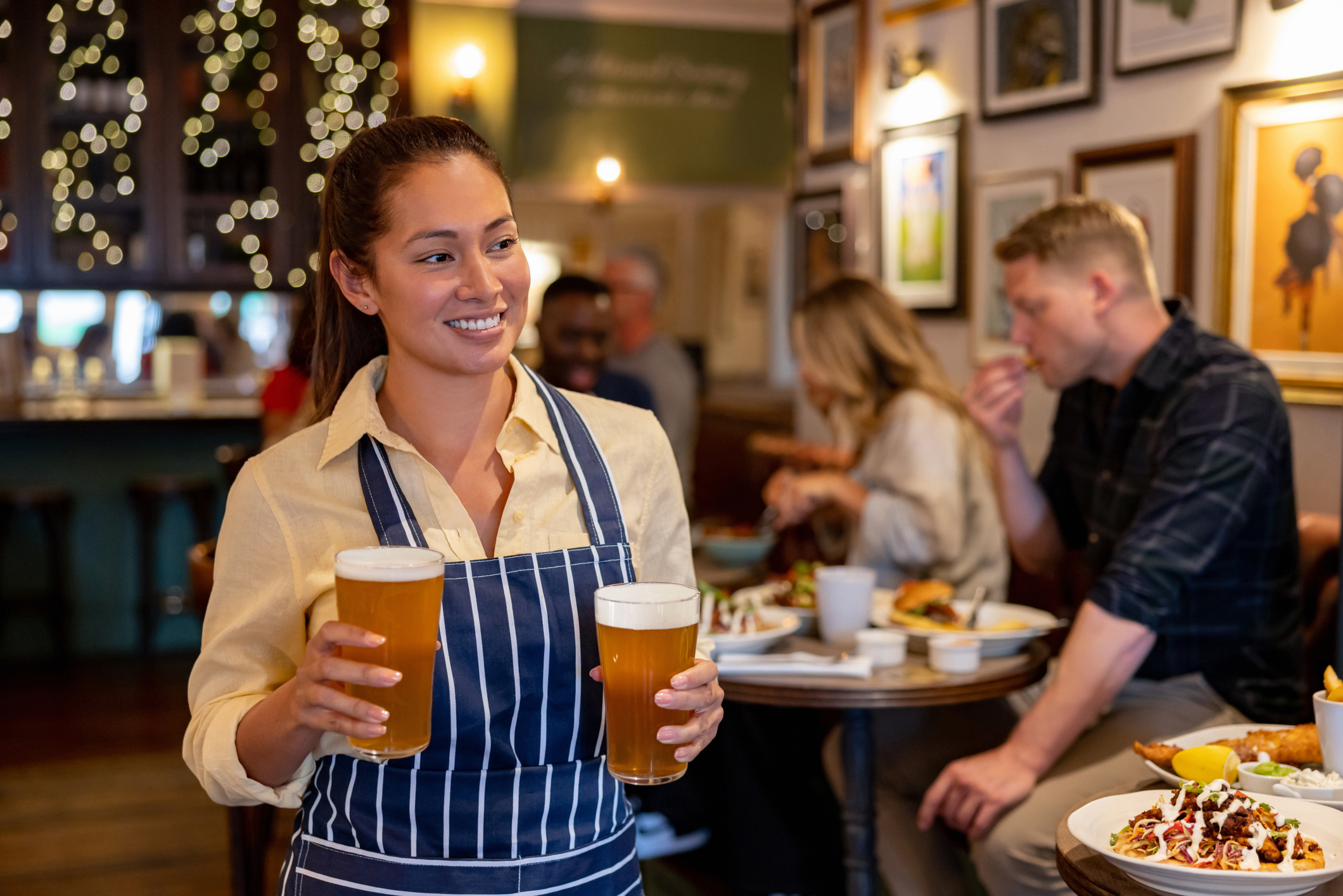 Waitress in striped apron serving two beers with patrons dining in background
