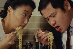 Two people are intensely eating noodles with chopsticks, one looking intently at the other.