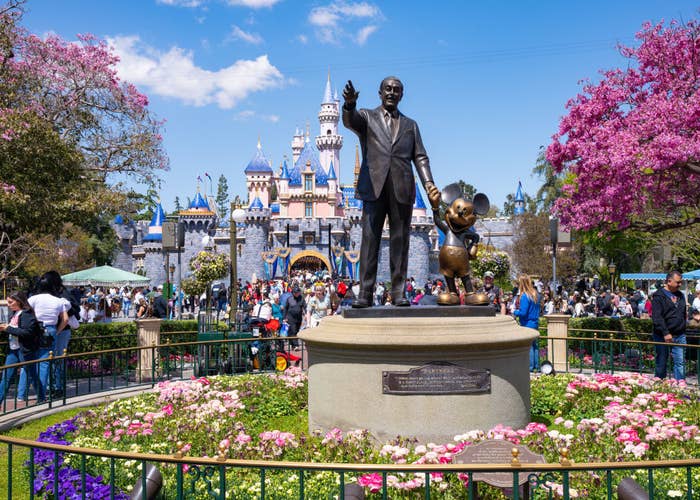 Statue of Walt Disney holding Mickey Mouse&#x27;s hand at Disneyland, with visitors and castle in background