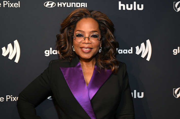 Oprah Winfrey posing with hands on hips, wearing a black suit with a purple collar detail, at a media event