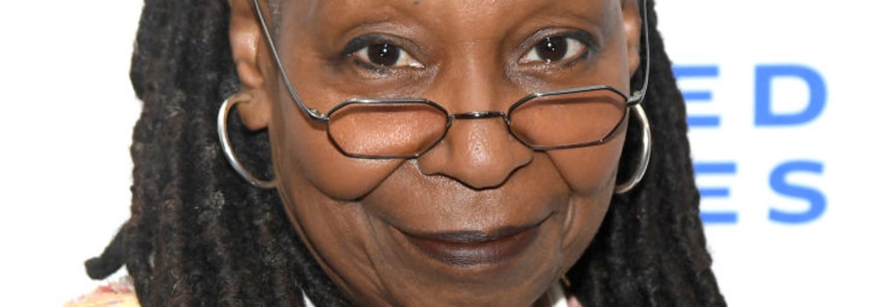 Whoopi Goldberg poses at an event, wearing glasses and a patterned jacket