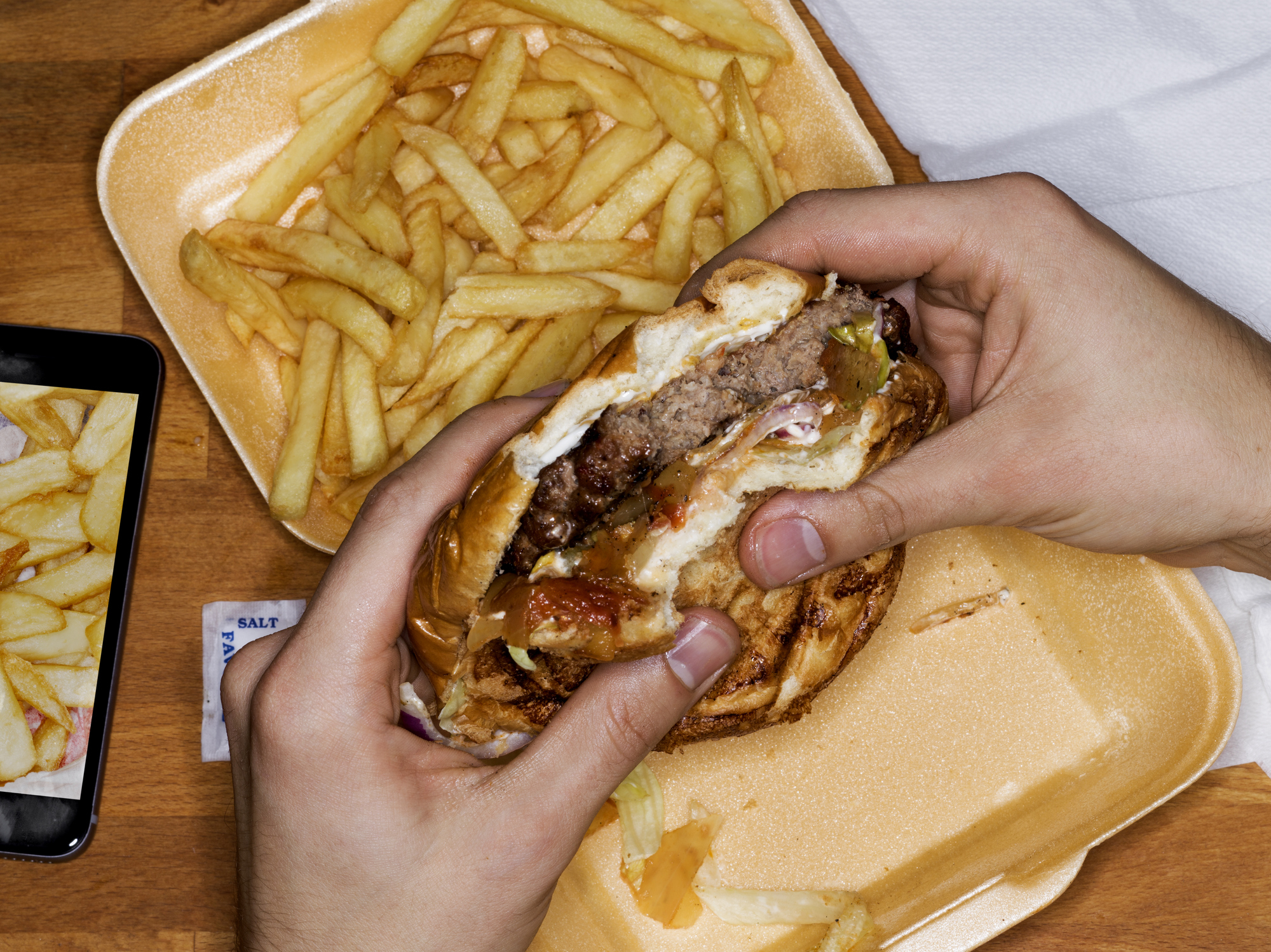 Person holding a half-eaten burger with fries on the side, on a tray, next to a smartphone