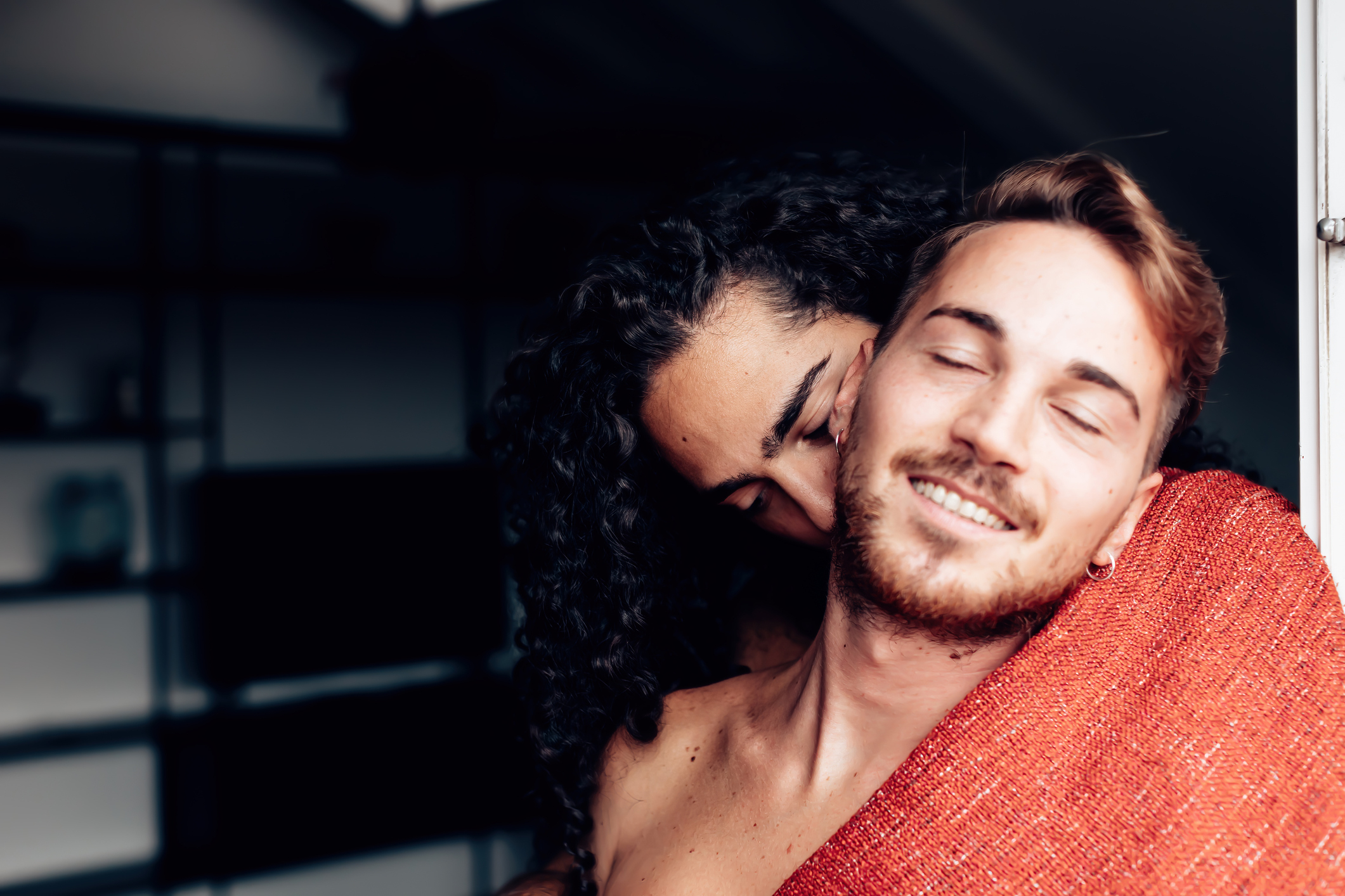 Two people embracing, one kisses the other's cheek, both smiling with closed eyes