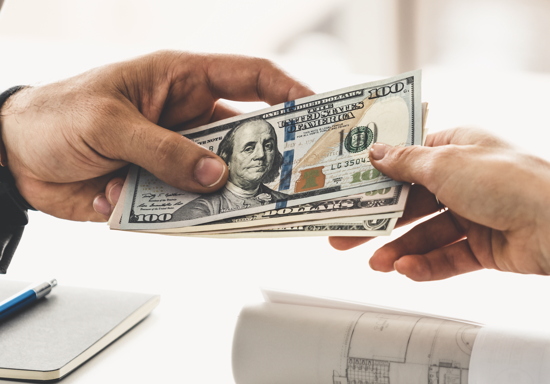 Person handing over U.S. dollar bills to another's outstretched hand indoors, suggesting transaction or payment