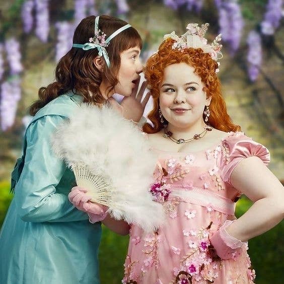 Dos personajes de &quot;Bridgerton&quot; en vestidos de época, uno con un abanico, en un fondo con vegetación