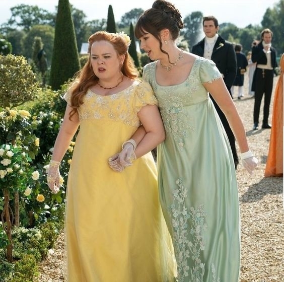 Dos personajes de Bridgerton en vestidos de época caminando y tomadas del brazo en un jardín