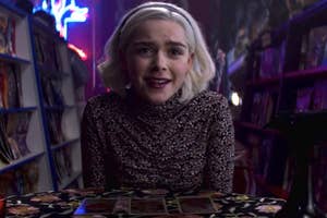 Image of Sabrina from 'Chilling Adventures of Sabrina' sitting at a table with tarot cards spread out. She wears a patterned top