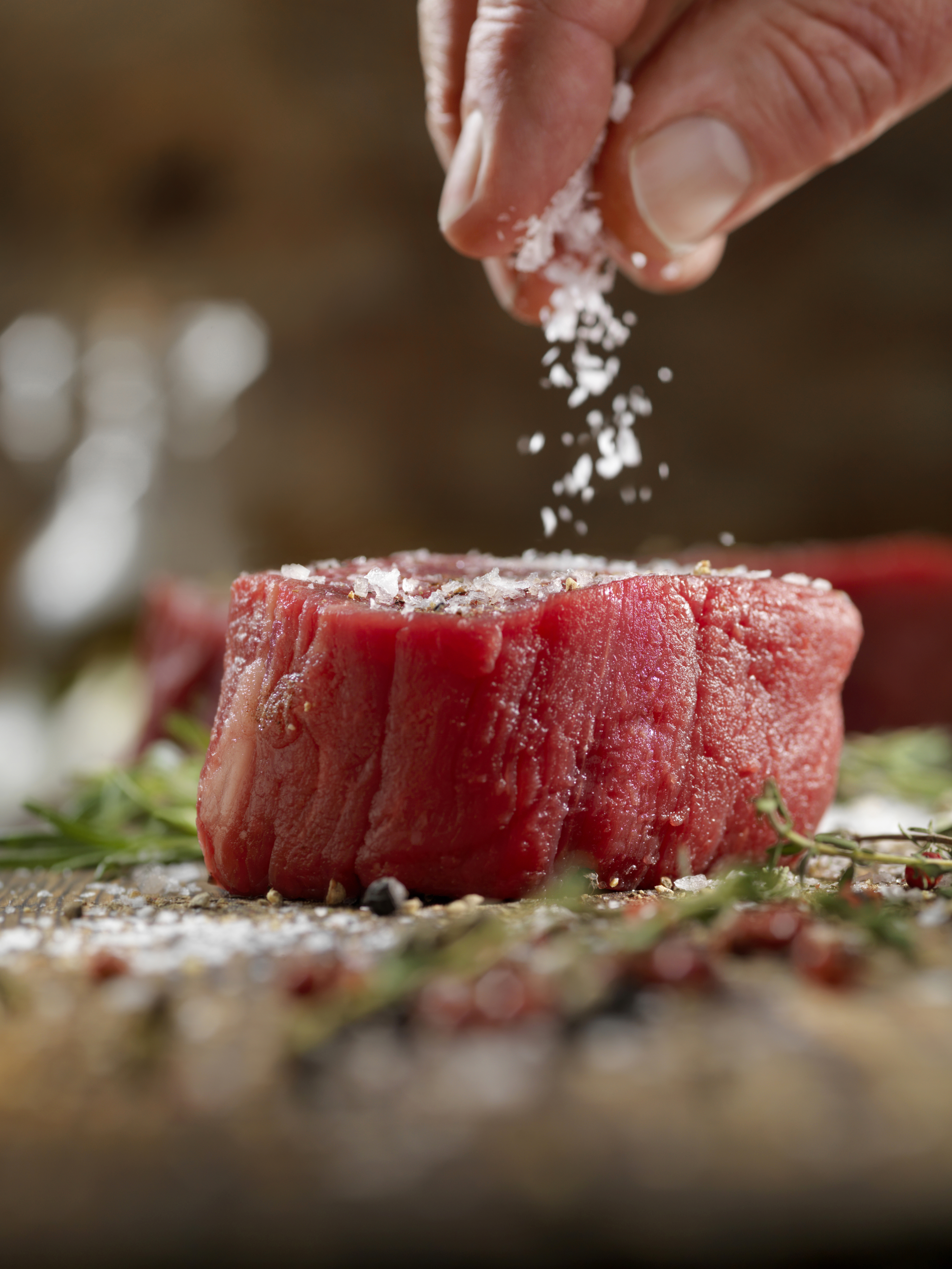 Hand seasoning a raw steak with salt on a wooden surface, surrounded by herbs