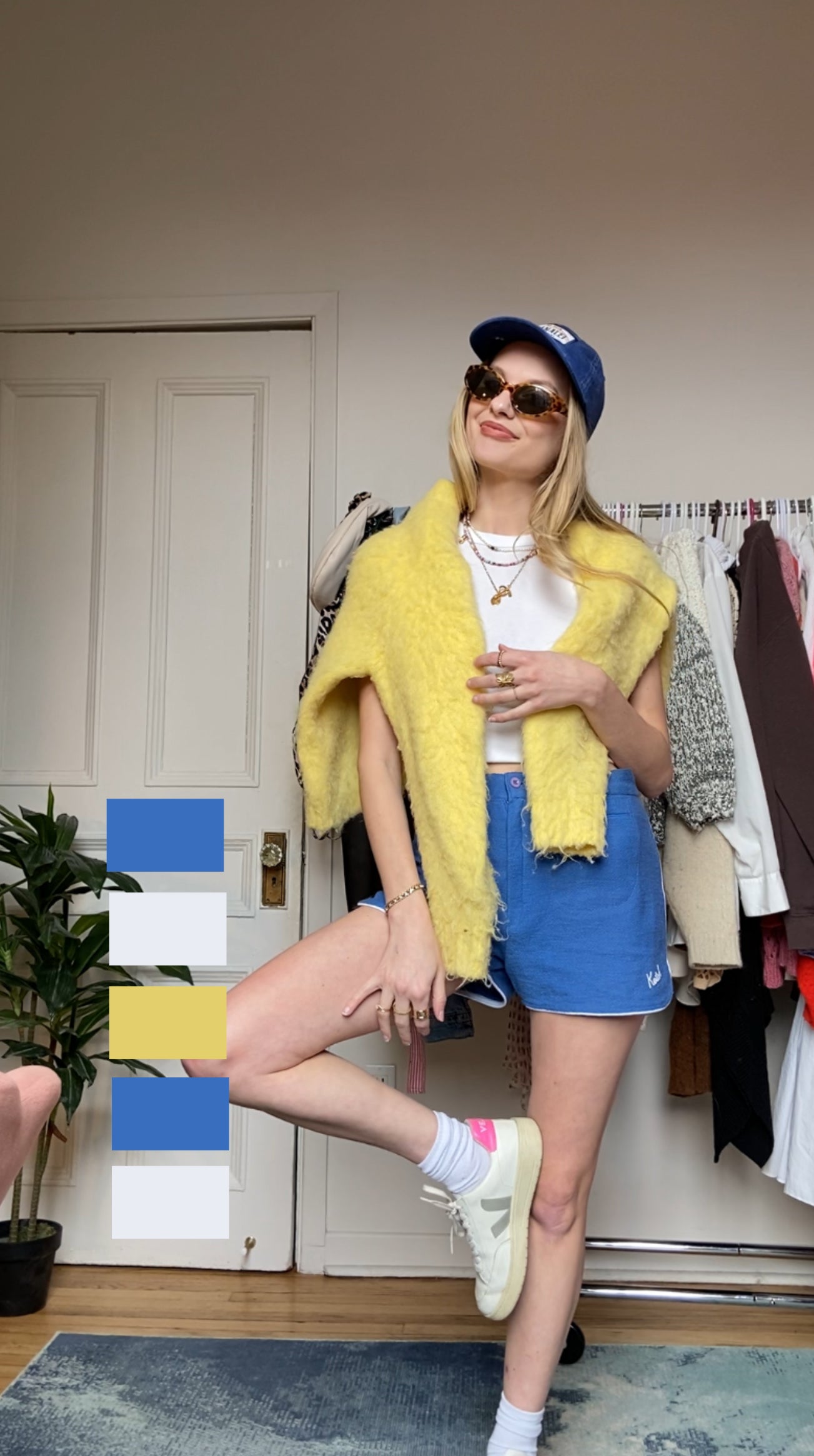 Woman in sunglasses and cap stands posing with one leg raised, wearing a yellow furry jacket and denim shorts