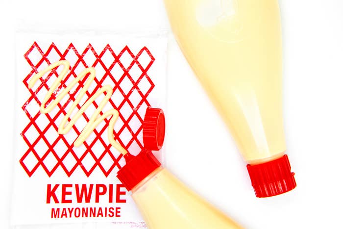 Dua botol mayones Kewpie tergeletak diagonal dengan tutup menghadap ke bawah, dengan latar belakang putih dekat logo merah