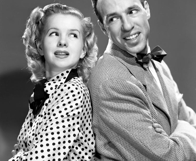 Two vintage Hollywood stars, the man in a tweed suit and bow tie, the woman in a polka dot dress with a bow collar