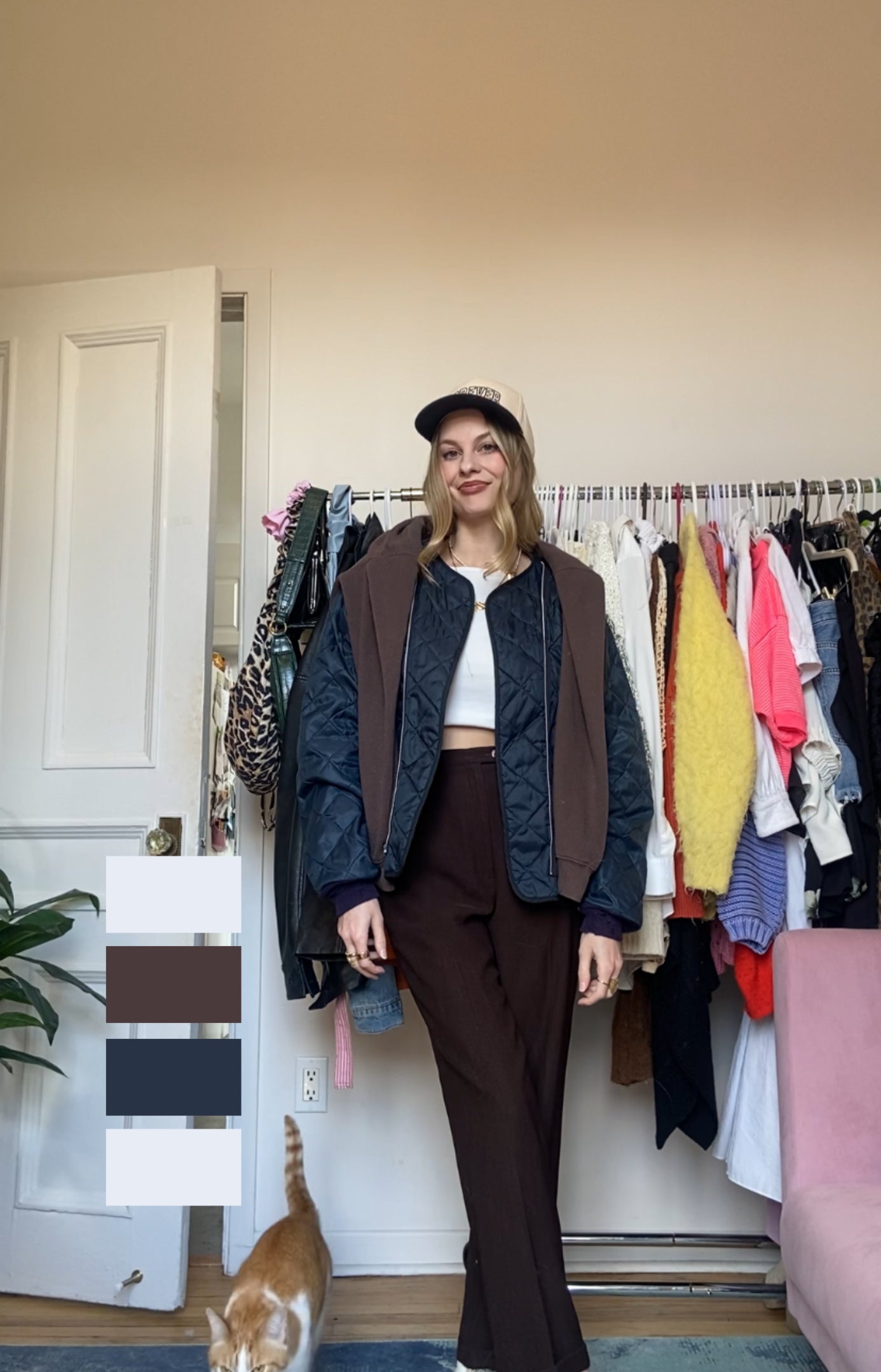 Person in a layered outfit with a cap stands in a room with clothes rack and a cat walking by