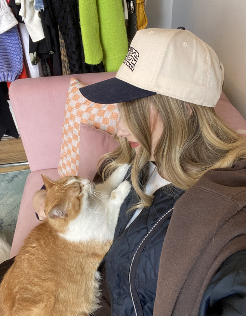 Person in a cap and jacket cuddling with an orange cat on a pink chair