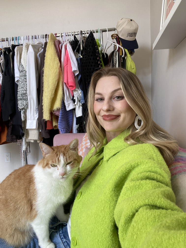 Woman in green jacket posing with an orange and white cat, with clothing rack in background