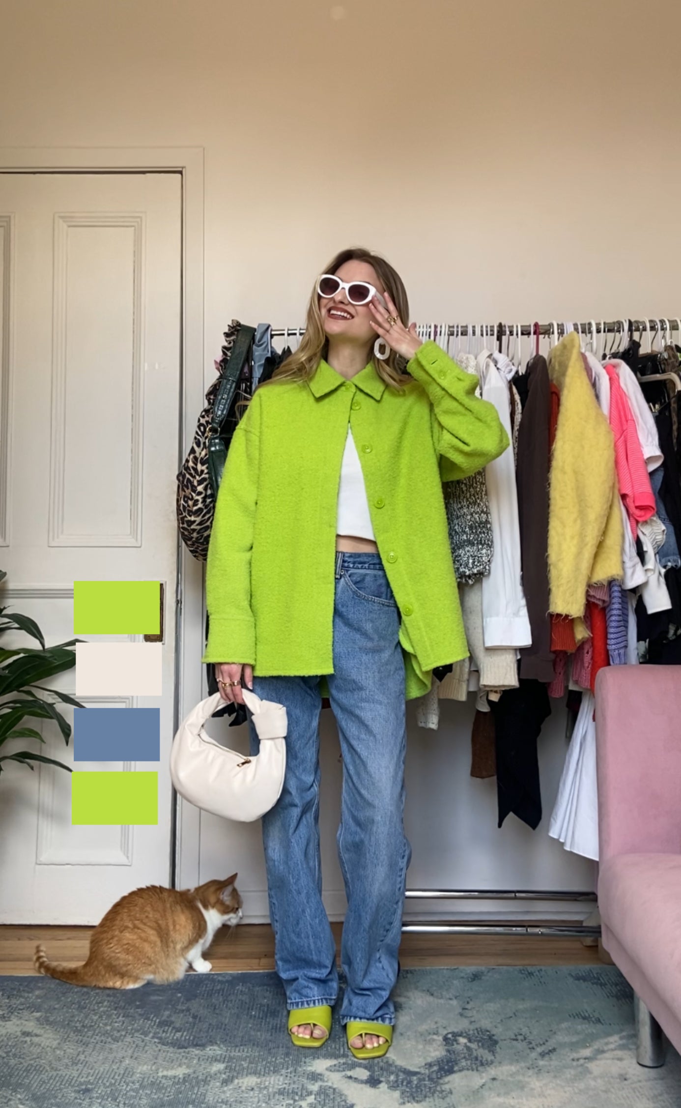 Woman in a bright green blazer and jeans with sunglasses, holding a white bag, standing by a rack of clothes with a cat nearby