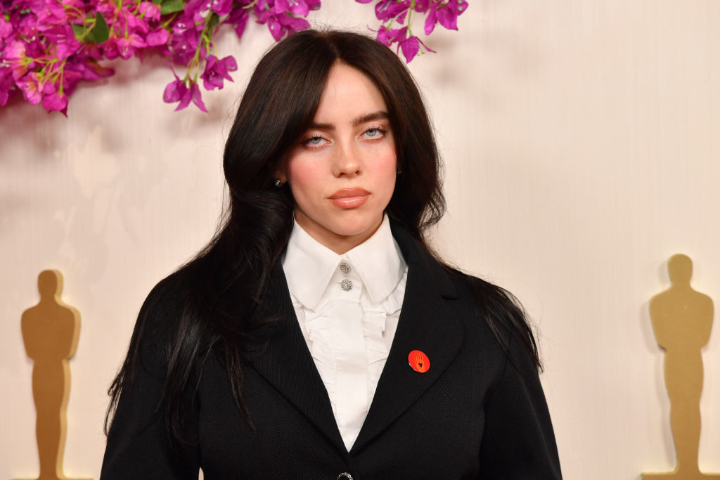 Billie Eilish in a shirt and blazer at an event with Oscar statues in the background