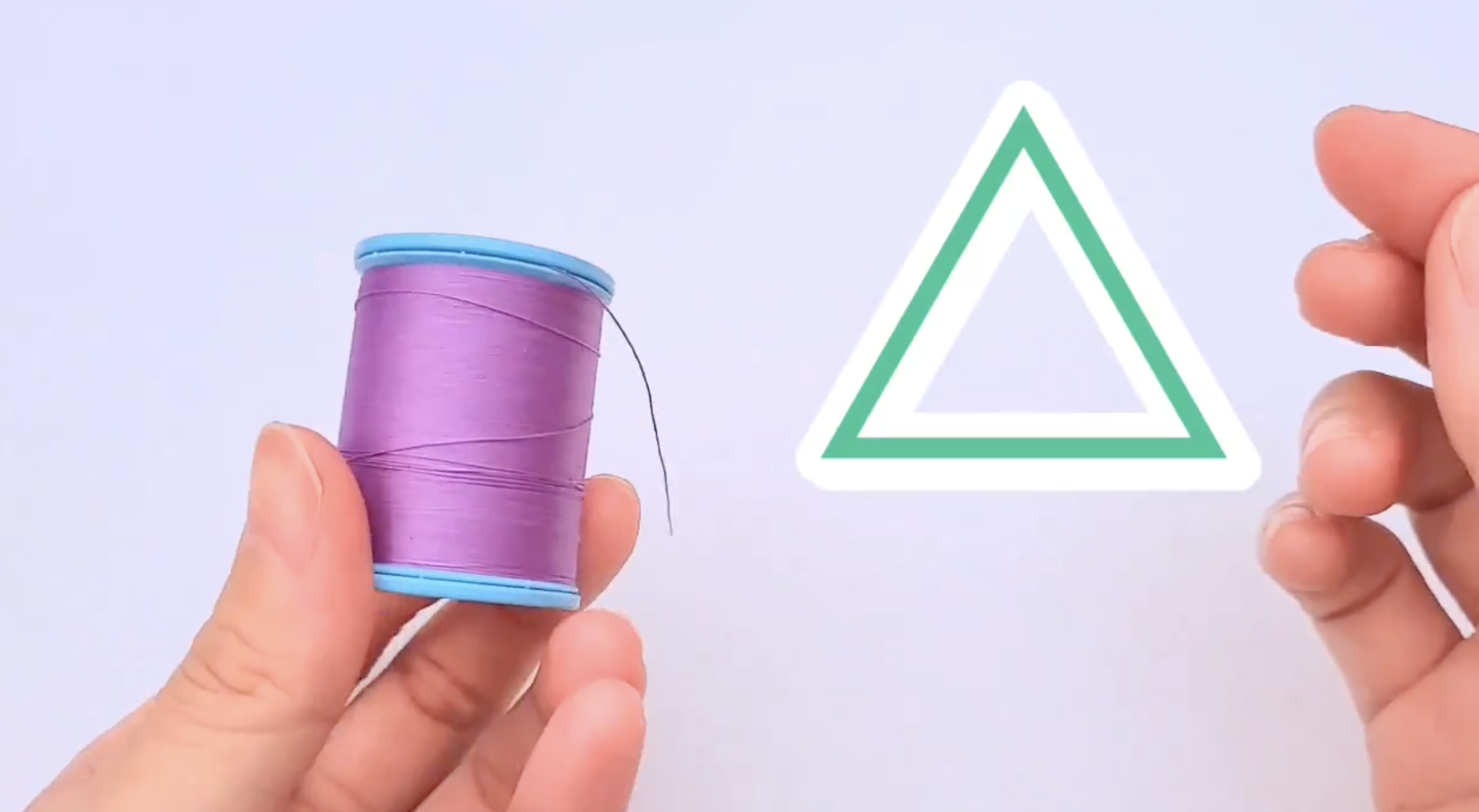 手が紫の糸が巻かれた糸巻きを持っており、隣には緑色の三角形の形の紙が浮かんでいます。