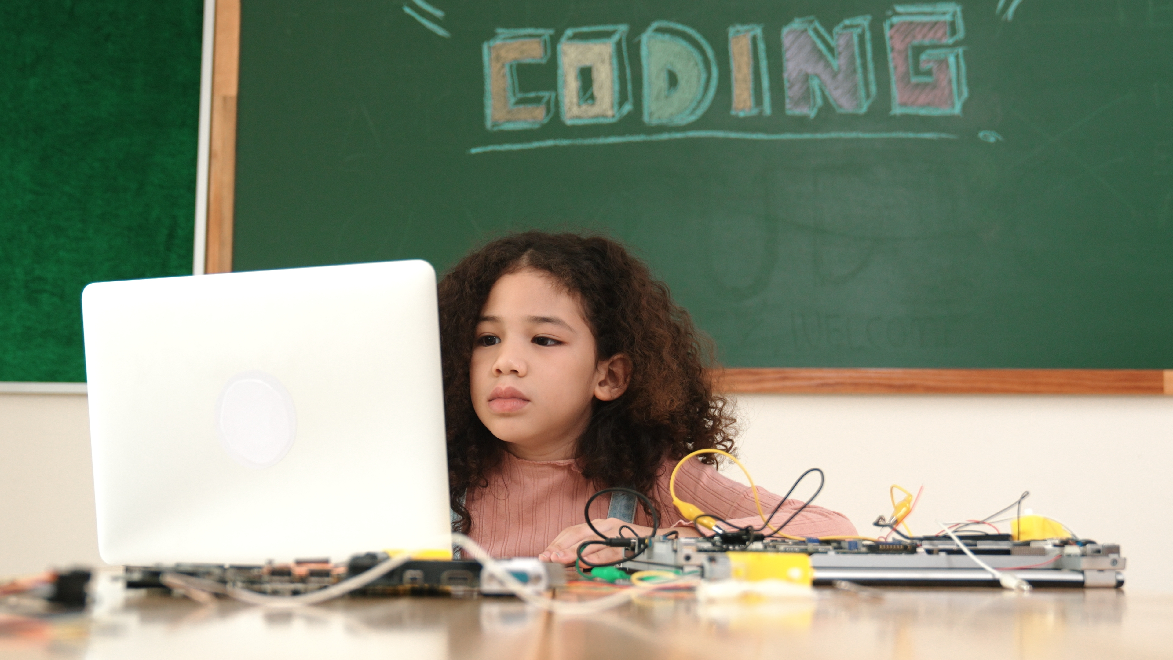 Young girl at a desk with a laptop and electronic components, with &#x27;CODING&#x27; written on the chalkboard behind her