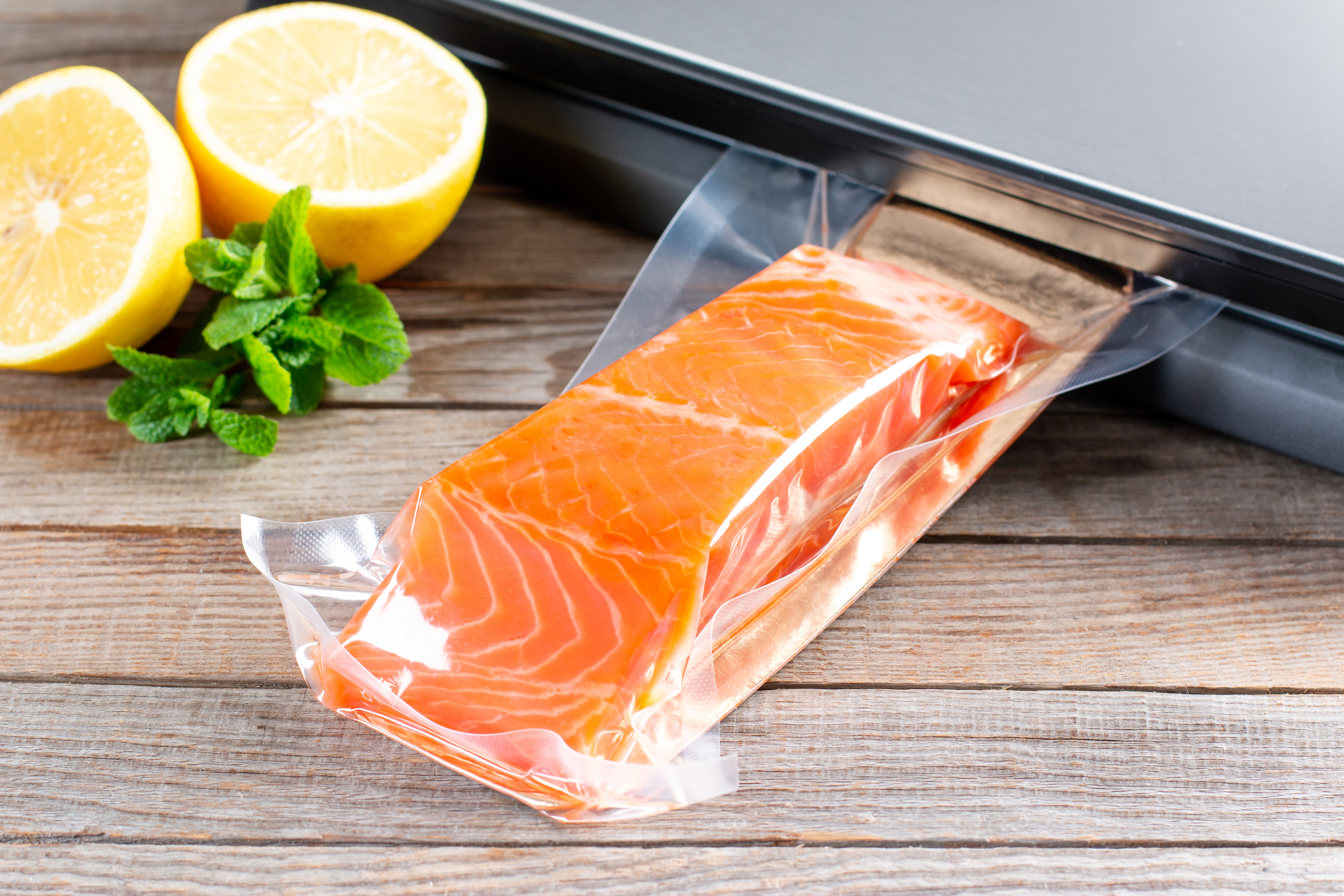 Vacuum-sealed salmon beside lemon halves and mint on a wooden surface, with a laptop