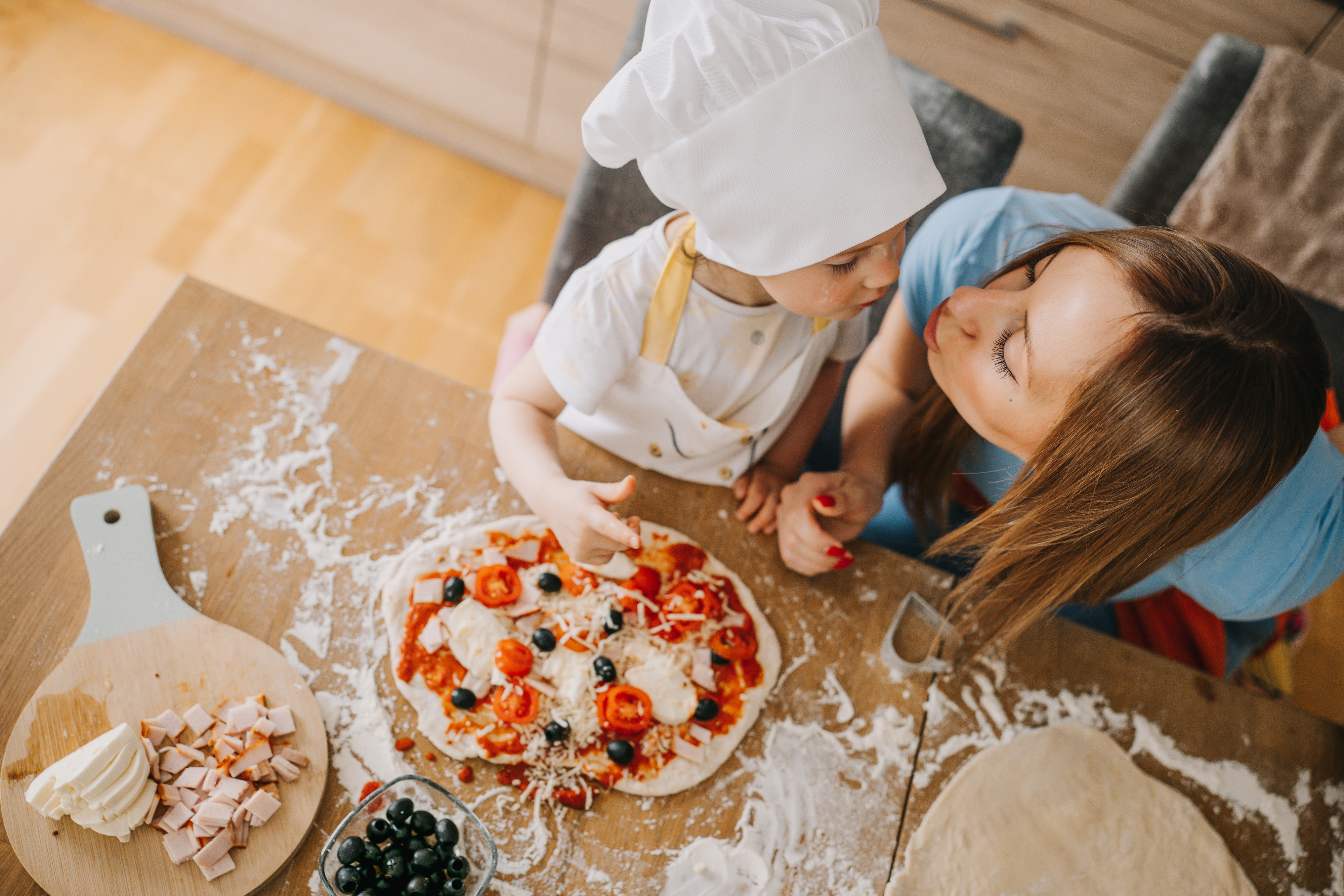 Adult and child in chef hats making pizza together on a wooden table