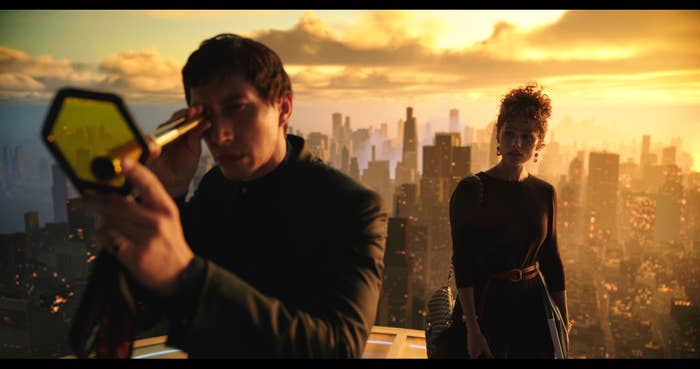Two characters from a film, one holding an object, with a cityscape in the background