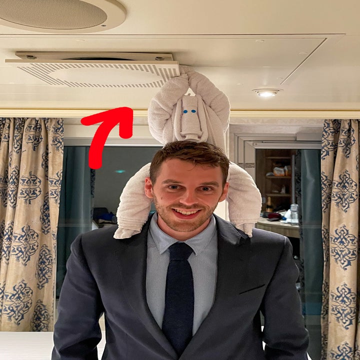 Man smiling with a towel folded like an elephant on his head, in a suit, indoors with curtains behind