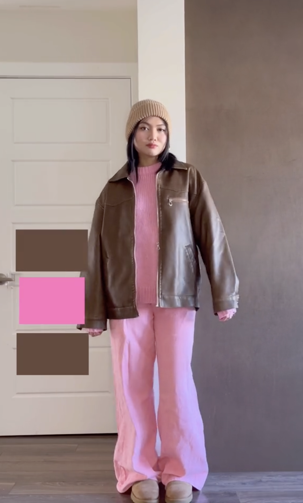 Woman in oversized jacket and wide-leg pants stands indoors, hands slightly extended. A pink square obscures part of the image