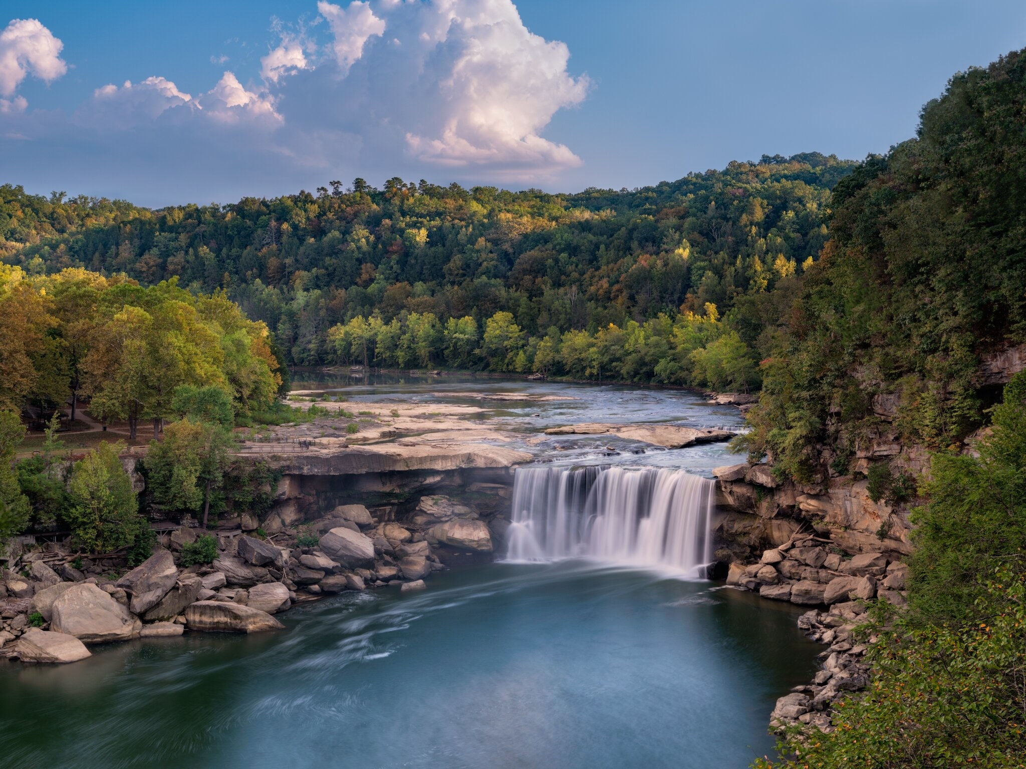 Waterfall cascading into a river surrounded by forested cliffs under a cloudy sky