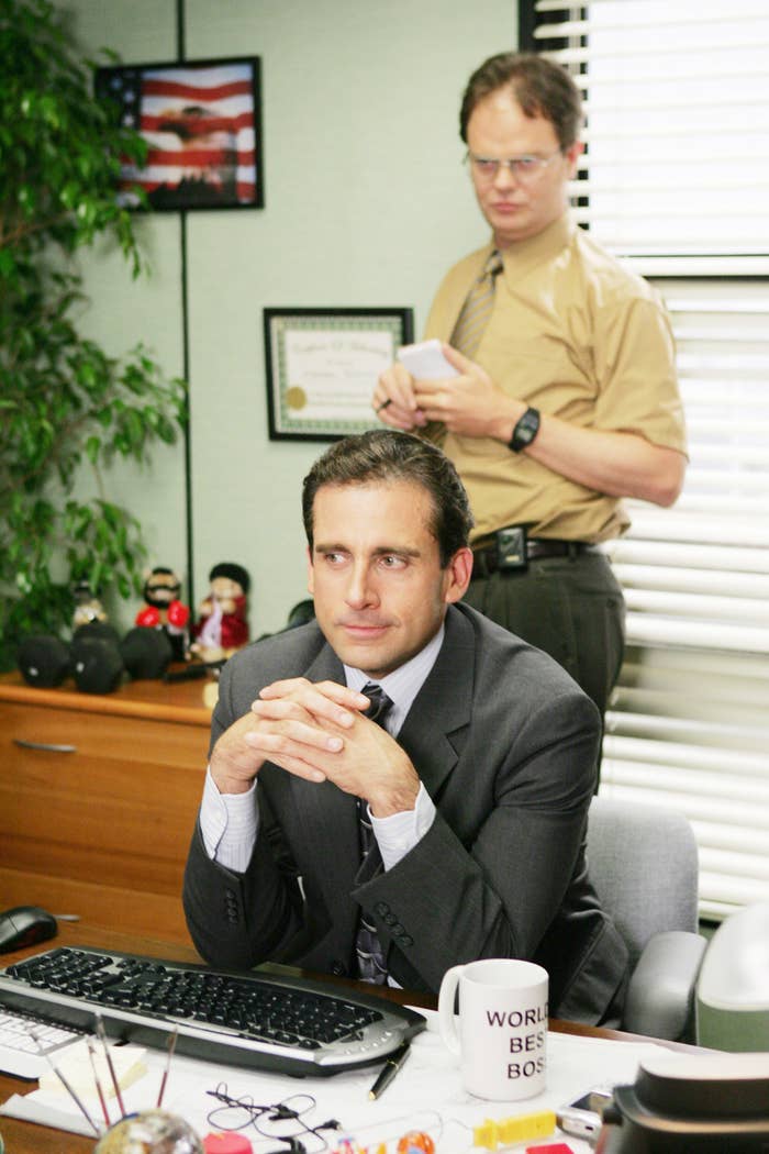 Michael Scott sits at his office desk while Dwight Schrute stands behind in the scene from The Office