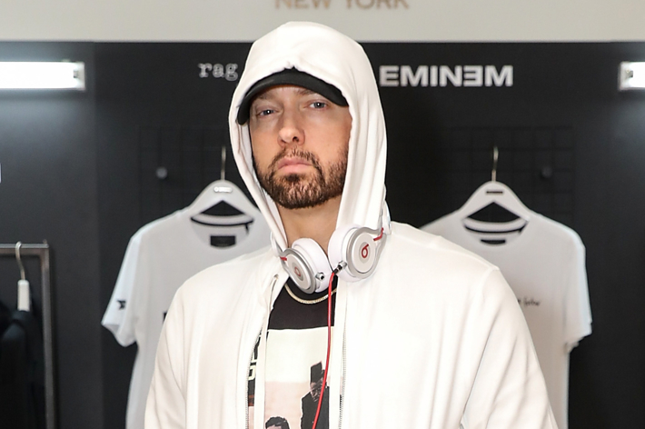 Eminem standing in a store, wearing a white hoodie, headphones around neck, expressionless