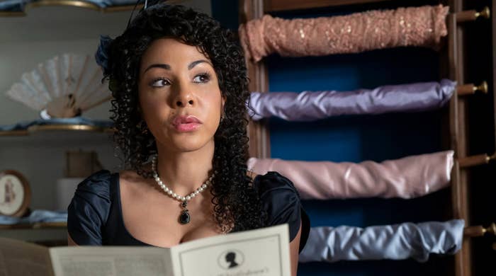 Mujer con vestido de época mira hacia arriba, rodeada de telas, en escena de serie de TV