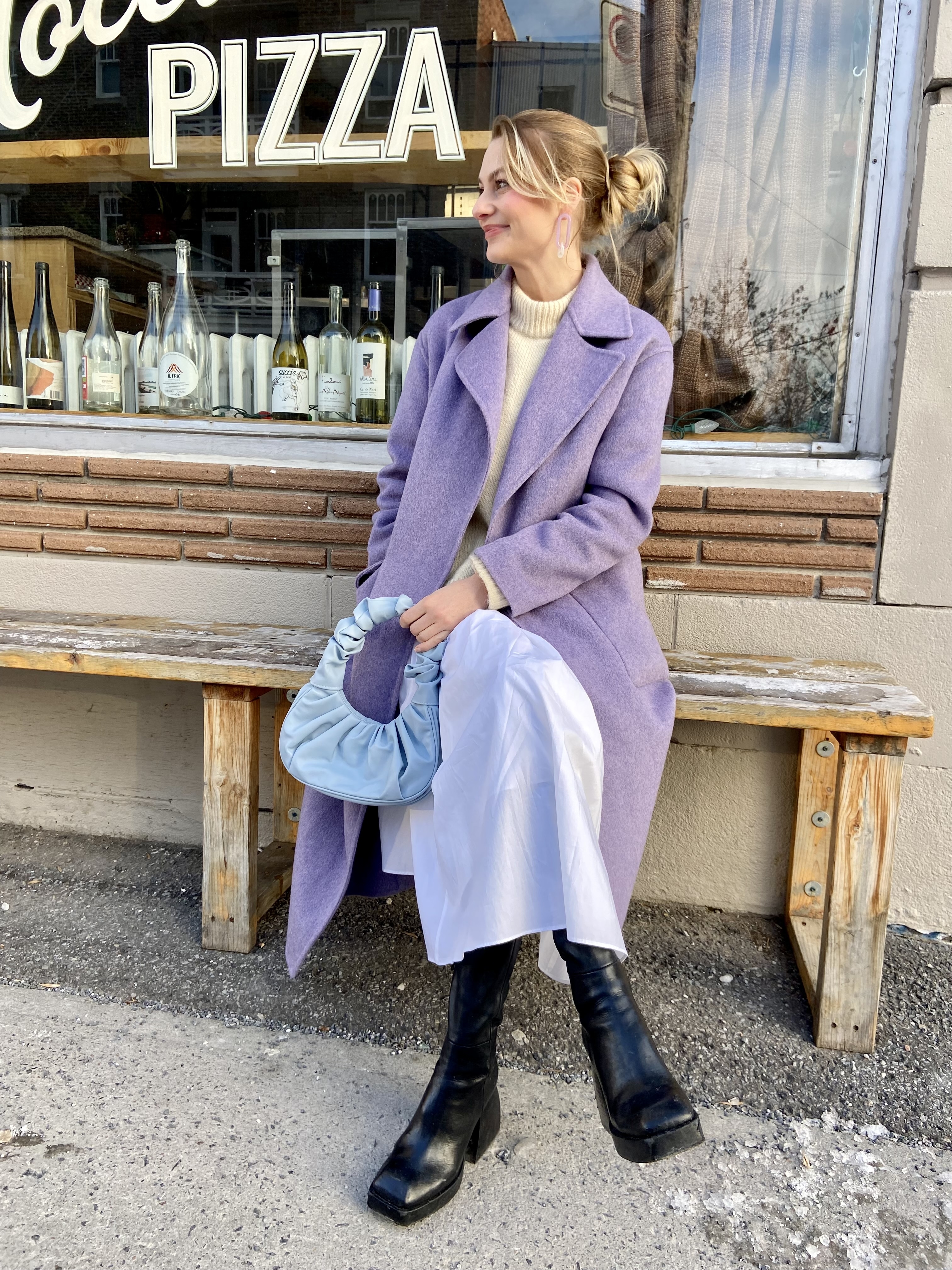 The author sitting on bench outside PIZZA shop in purple coat, white dress, black boots, holding blue bag