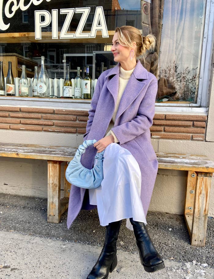 The author sitting on bench outside PIZZA shop in purple coat, white dress, black boots, holding blue bag