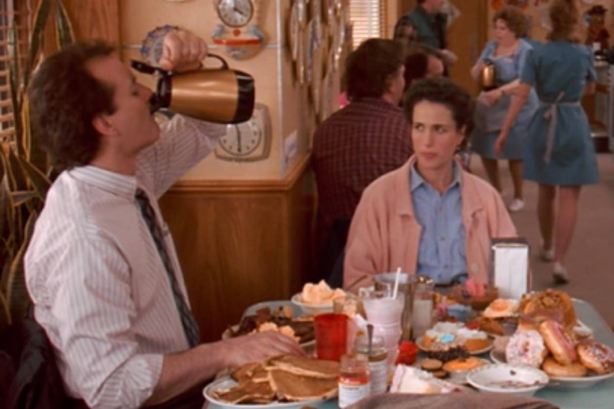 Man chugging syrup beside woman at diner table with excessive breakfast foods