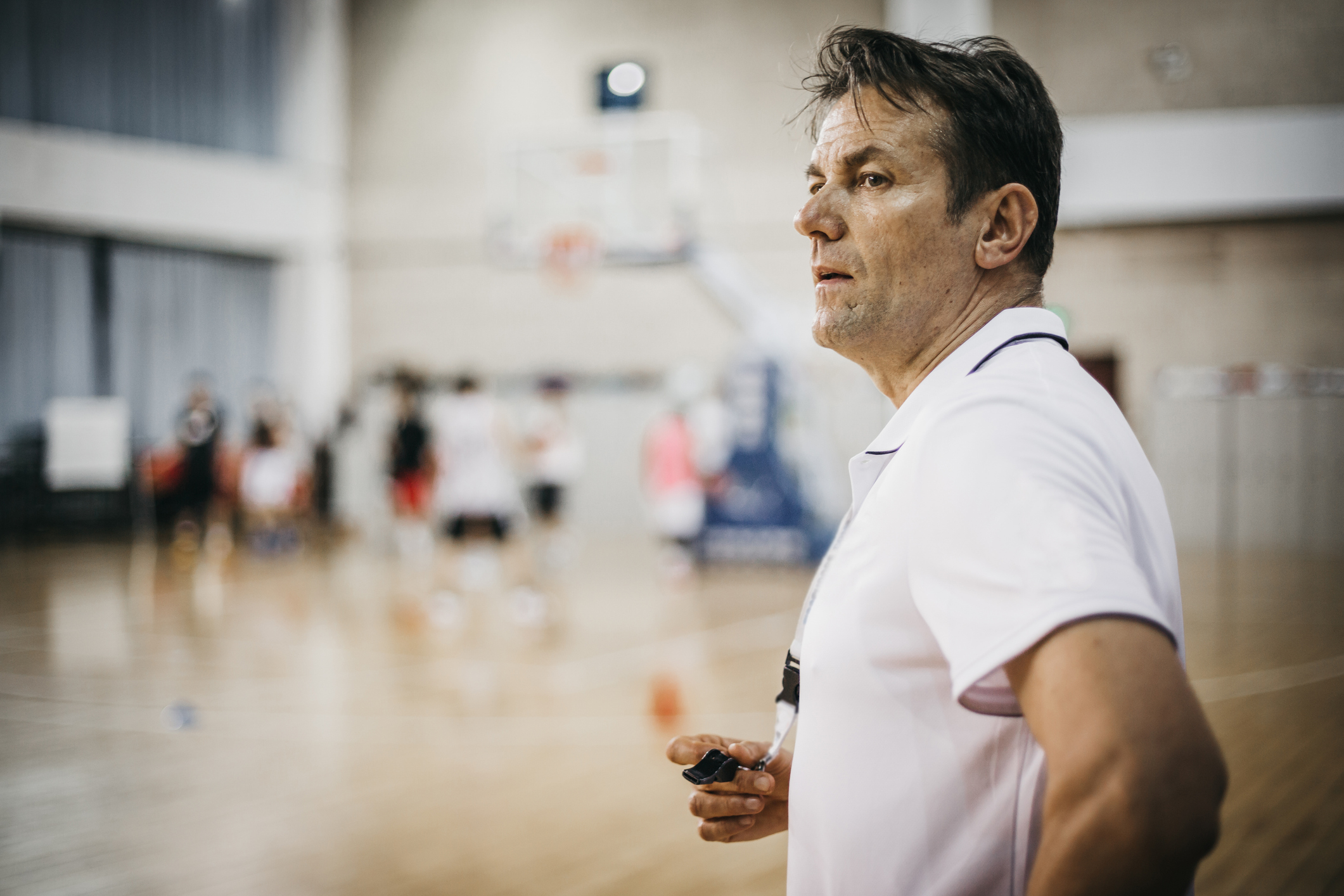 Coach observing players in a gym, holding a whistle, focused on team's performance