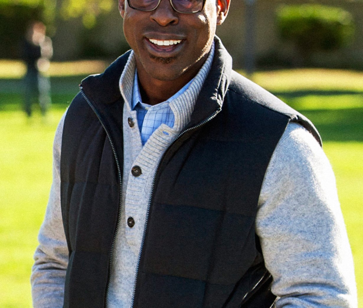 A man in glasses smiles wearing a layered sweater and vest, standing outside