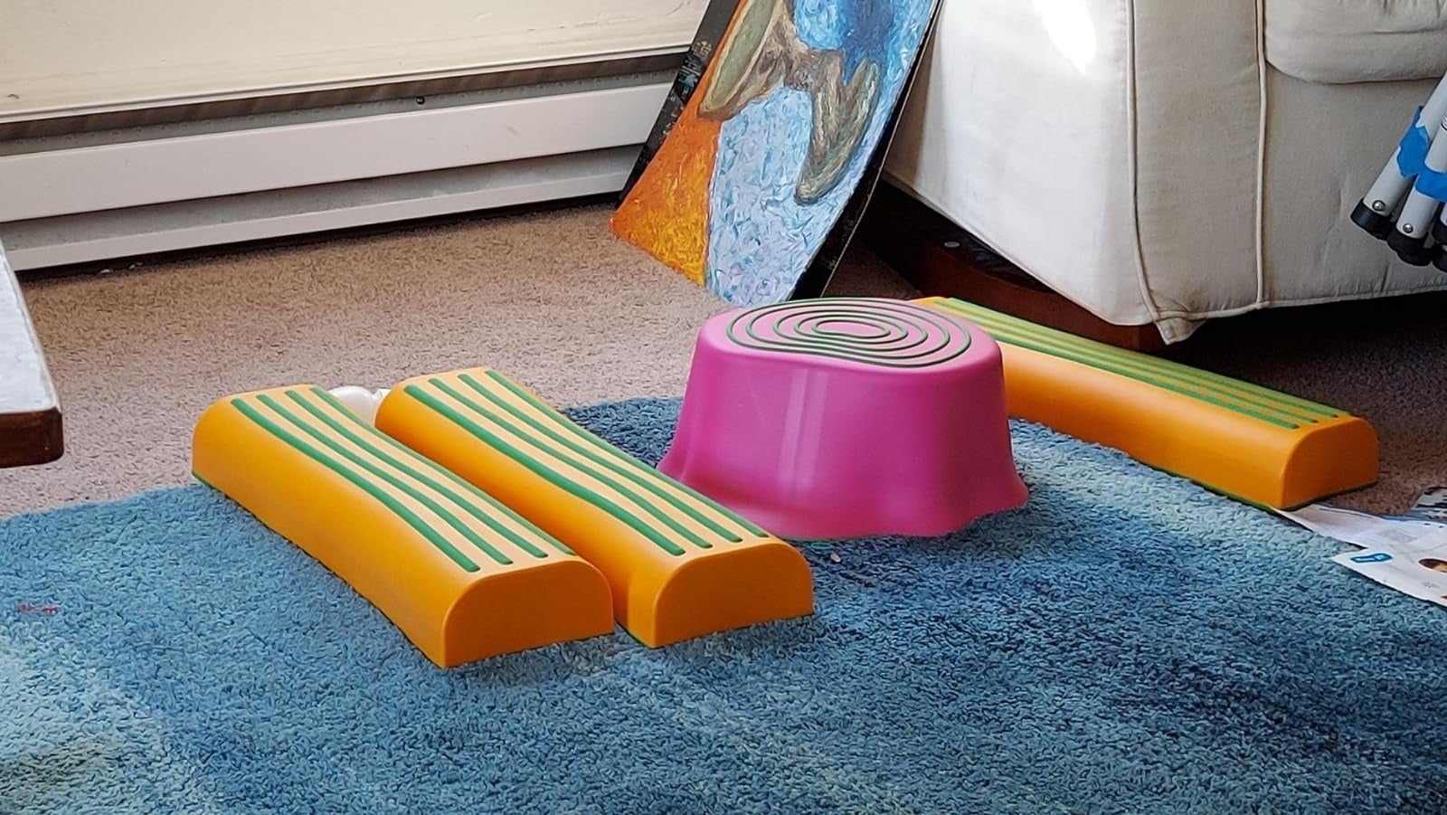 Plastic equilibrium  beams and a pinkish  stepping stool arranged connected  a bluish  carpet adjacent   a couch, adjacent  to a leaning painting
