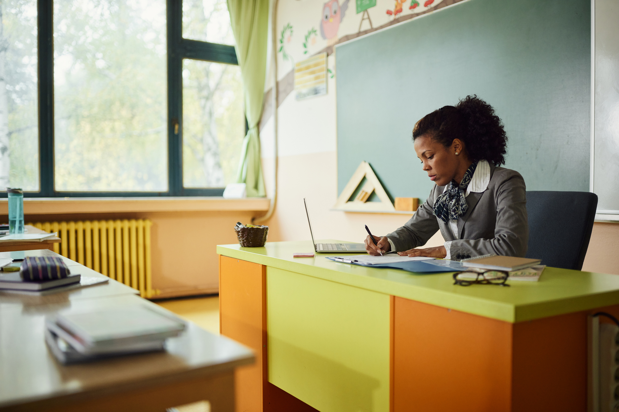 A teacher is working at her desk in a classroom, reviewing documents and using a laptop