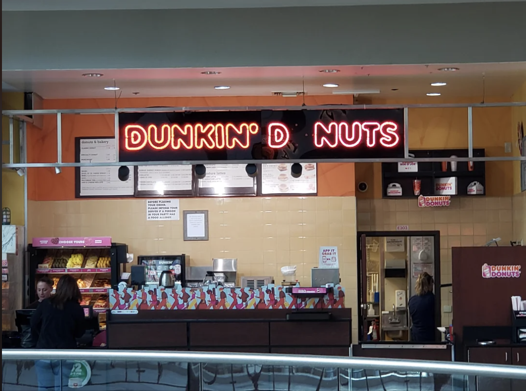 Sign above a cafe reads "DUNKIN' D NUTS" due to missing letters, with customers and staff visible