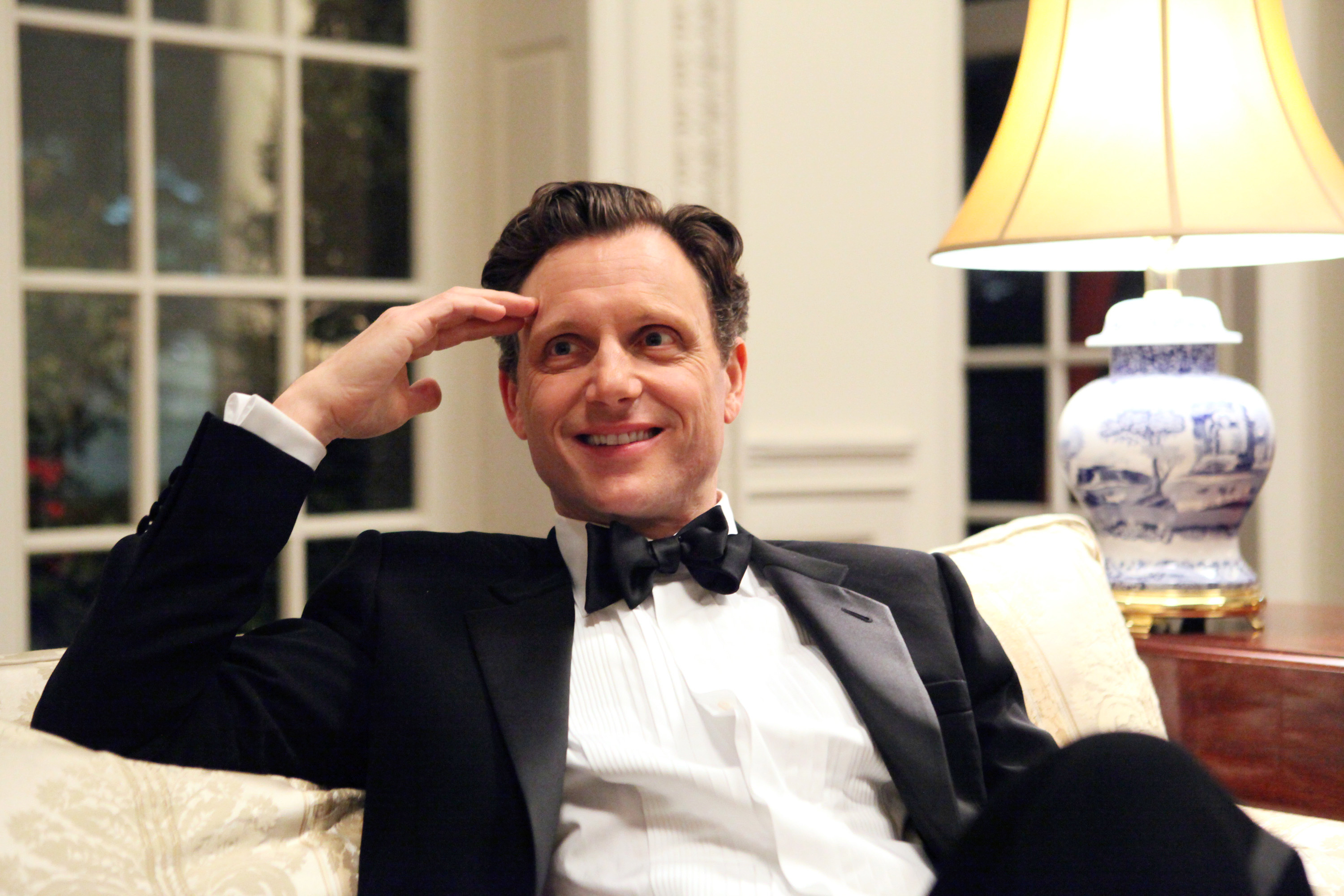 Man in a black tuxedo and bow tie saluting from a chair, apparently from a TV show or movie scene